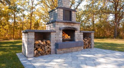 Quarry stone outdoor fireplace by Barkman Concrete, available at Ron Paul Garden Centre. Fire is lit in the fireplace and has cut wood on either side of it. Showcased on a stone patio.